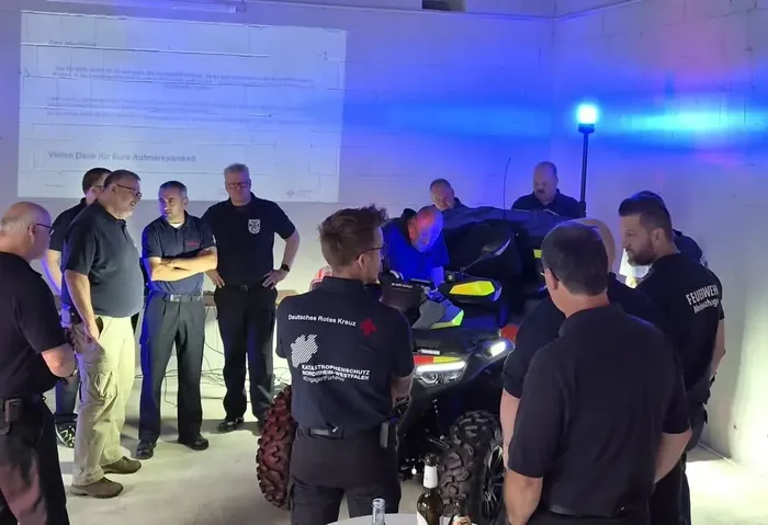 Einsatzkräfte versammeln sich in einem blau beleuchteten Raum um ein Quad; im Hintergrund ist eine Projektion mit einem Dankestext zu sehen.