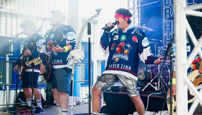Dr. Jochen Veit singt in einem blau-weißen Roosters-Trikot und silbernen Shorts bei einem Live-Auftritt auf einer Bühne.