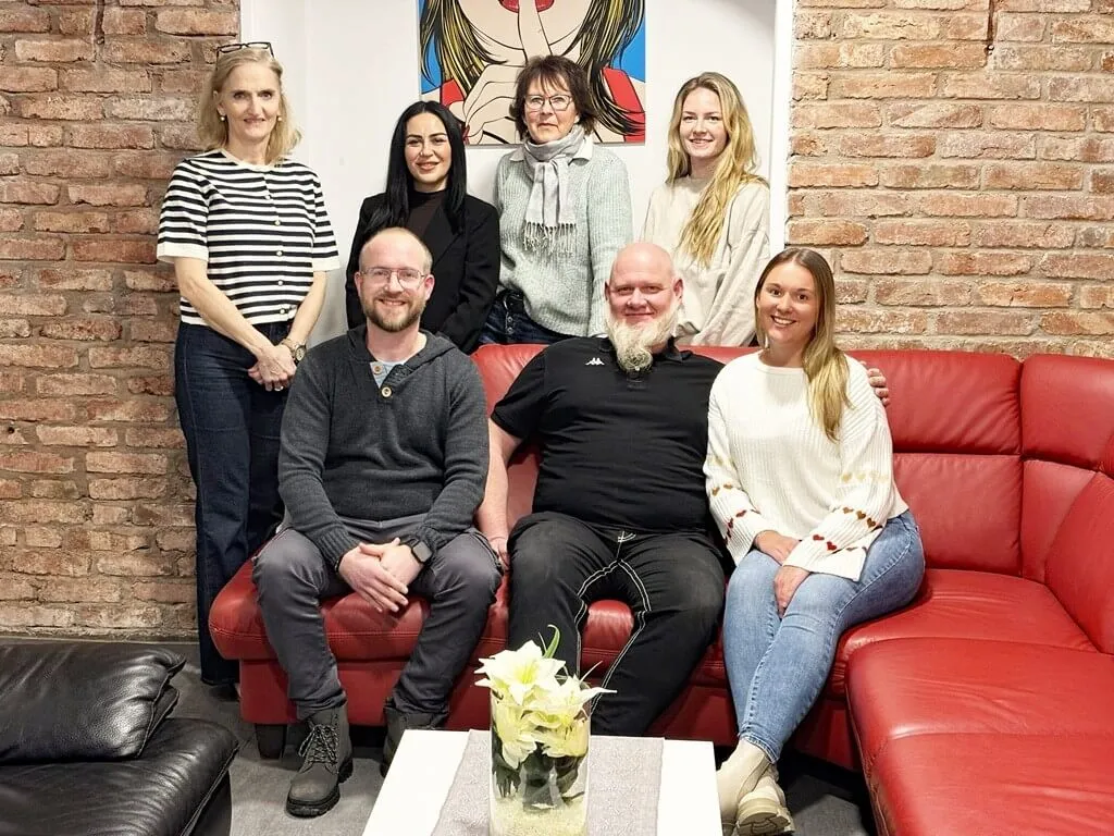 Sieben Personen sitzen oder stehen vor einer Backsteinwand und lächeln in die Kamera. Im Vordergrund sitzt ein Mann mit Bart auf einem roten Sofa, links und rechts von ihm jeweils zwei weitere Personen. Im Hintergrund stehen drei Frauen.