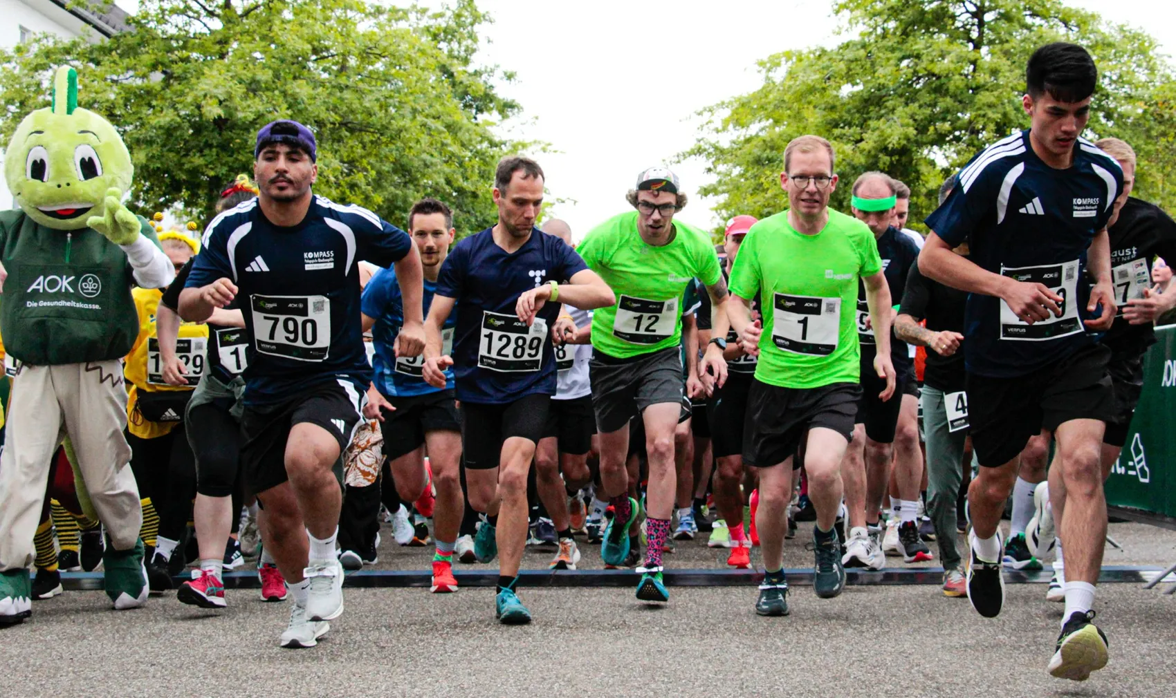 Läufer starten bei einem Lauf im Sauerlandpark, viele tragen Startnummern und Sportkleidung. Links begleitet eine große grüne Comicfigur die Läufer.