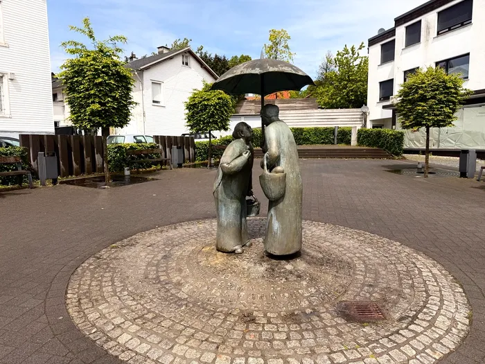 Bronzestatue eines Paares unter einem Schirm auf einem Platz in Halver, umgeben von einem kreisförmigen Pflastermuster.