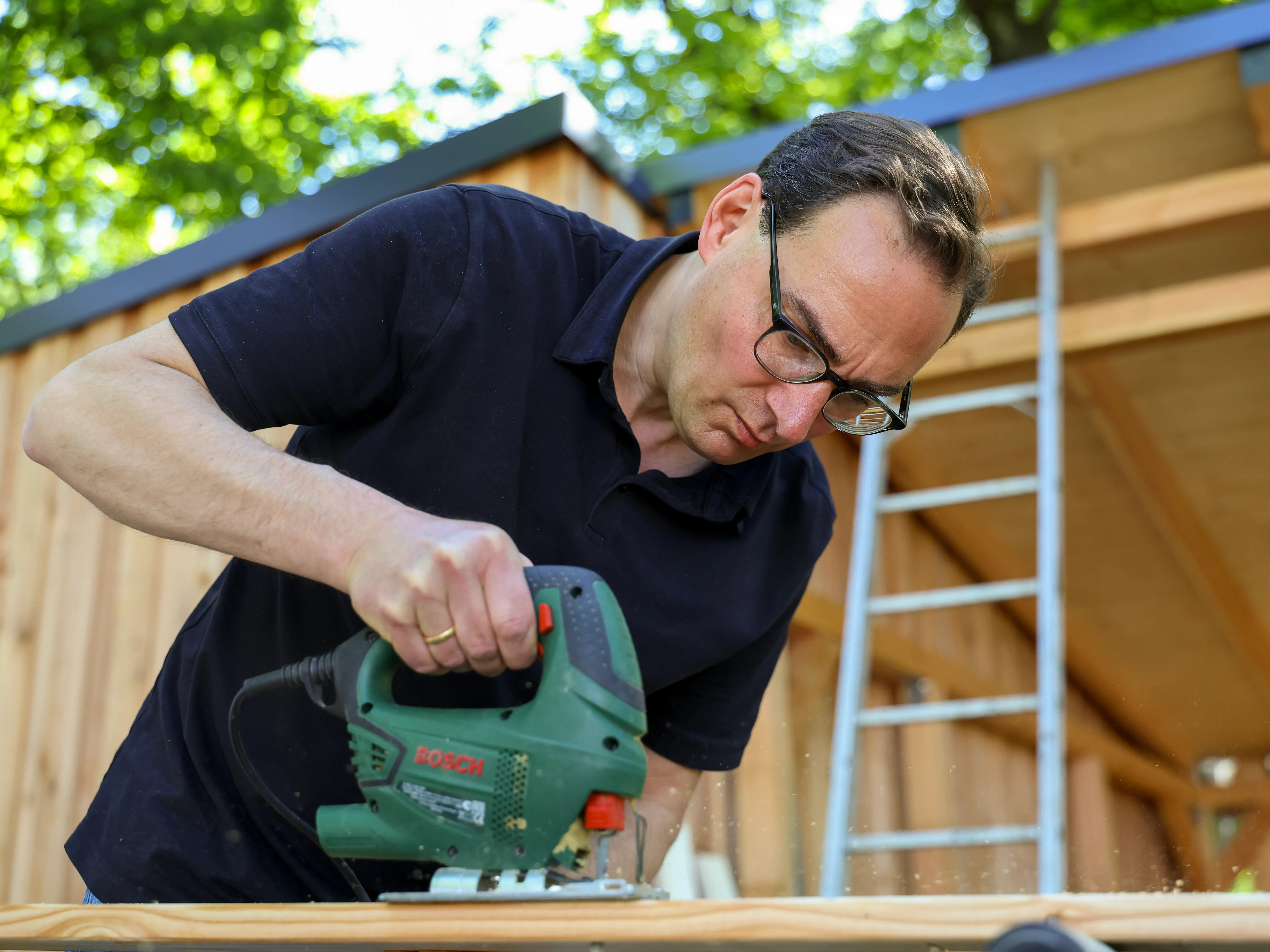 Ein Mann mit Schutzbrille und schwarzem T-Shirt sägt mit einer Stichsäge Holz vor einer Gartenhütte.