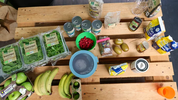 Eine Vogelperspektive zeigt eine Holztischoberfläche mit einer Auswahl an frischen Produkten und Lebensmitteln: Spinat, Bananen, Äpfel, Tomaten, Beeren, Hülsenfrüchte in Konserven, Sonnenblumenkerne, Reis und Chiasamen.