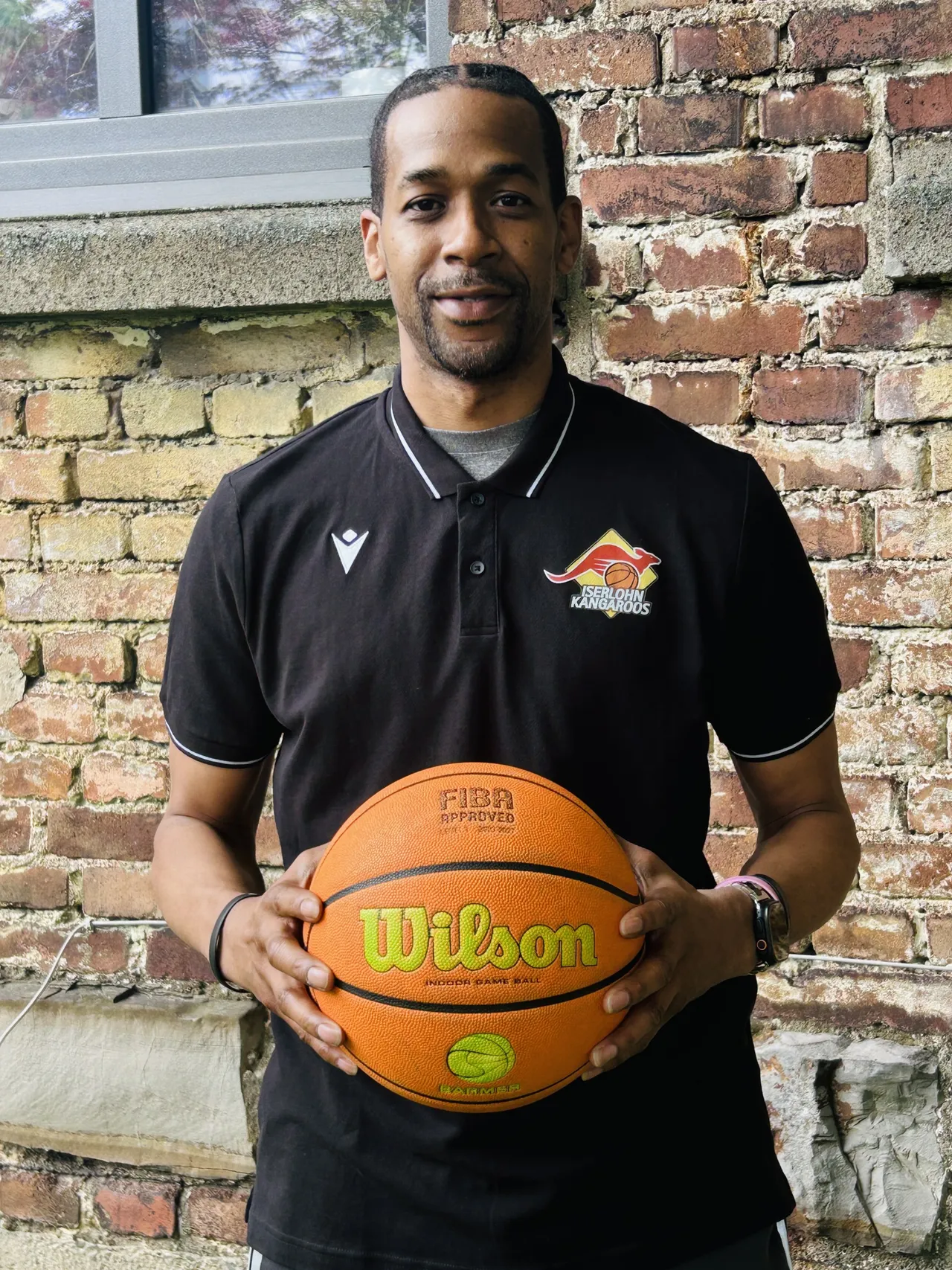 Gary Johnson, ein Mann mit dunkler Hautfarbe, hält einen Basketball mit dem Aufdruck Wilson und dem FIBA-Logo vor einer Backsteinmauer. Er trägt ein schwarzes Poloshirt mit dem Logo der Iserlohn Kangaroos.