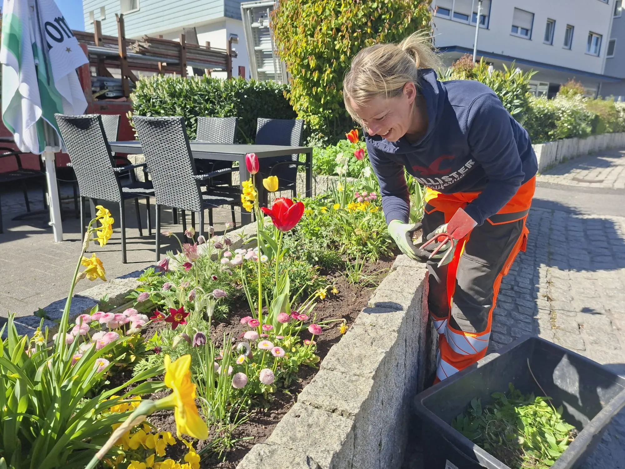 Frau gärtnert in einem Blumenbeet vor einem Café mit Terrassenmöbeln. Sie trägt eine blaue Jacke und orangefarbene Arbeitskleidung.