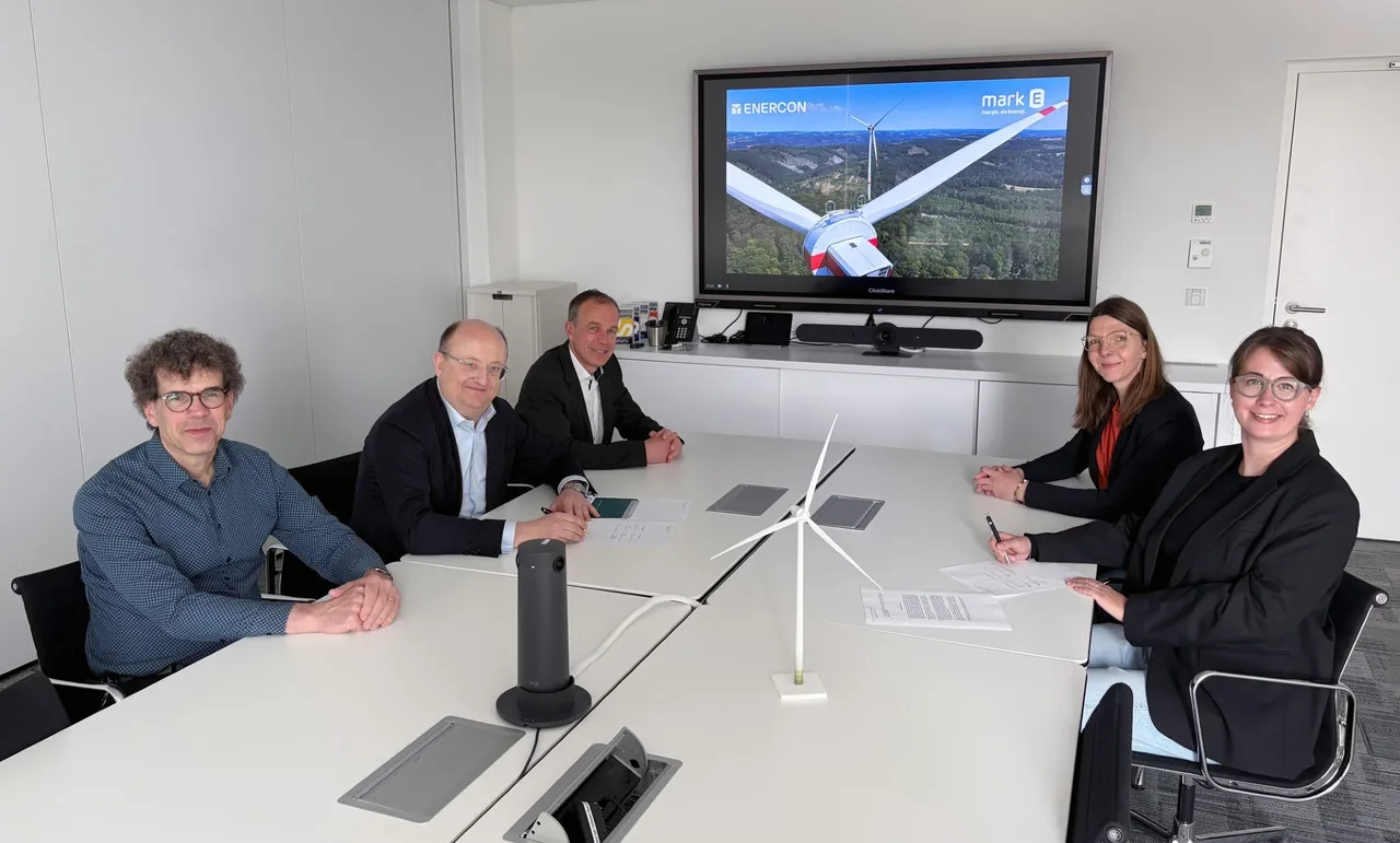 Fünf Personen sitzen an einem Konferenztisch und unterzeichnen Verträge. Im Hintergrund ist auf einem großen Bildschirm eine Windkraftanlage zu sehen, die das Projekt „Lüdenscheid-Kälberberg“ repräsentiert.