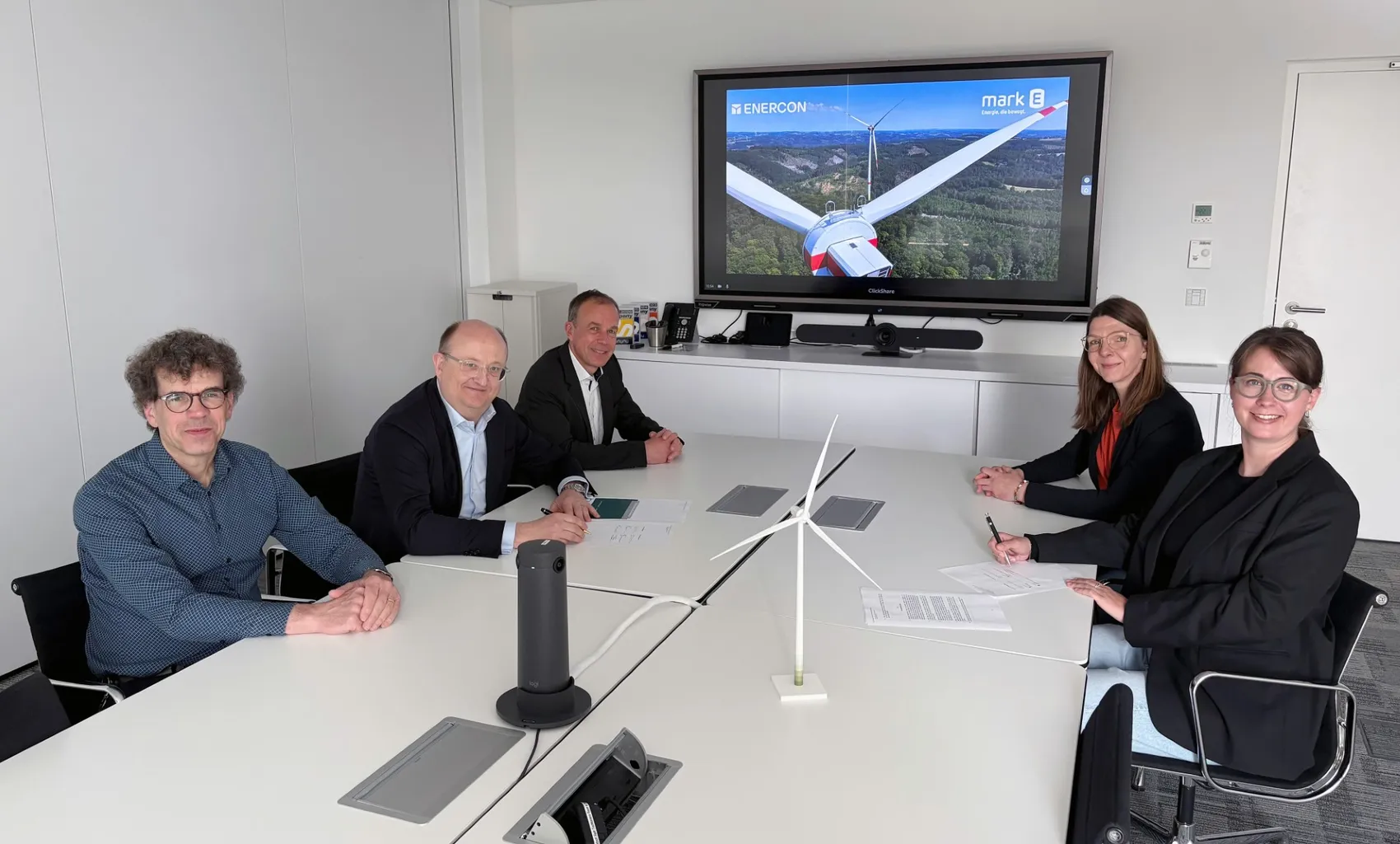 Fünf Personen sitzen an einem Konferenztisch und unterzeichnen Verträge. Im Hintergrund ist auf einem großen Bildschirm eine Windkraftanlage zu sehen, die das Projekt „Lüdenscheid-Kälberberg“ repräsentiert.
