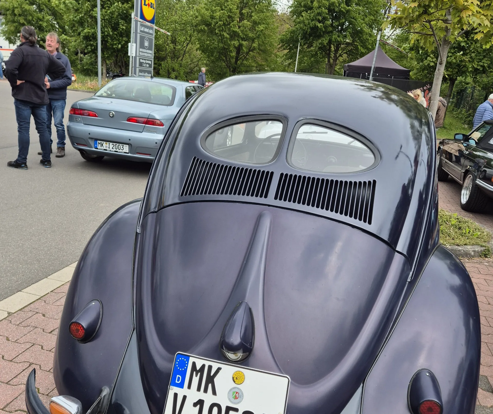 Heckansicht eines dunkelblauen VW Käfers mit Kennzeichen, im Hintergrund ein älterer silbergrauer Wagen und zwei Männer im Gespräch.