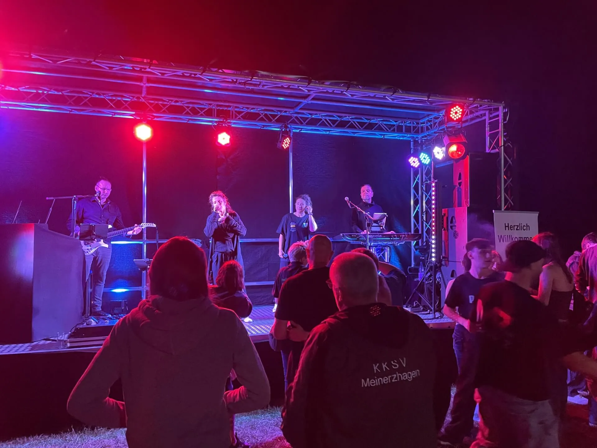 Eine Band spielt auf einer beleuchteten Bühne vor Publikum. Im Hintergrund ein Banner mit der Aufschrift „Herzlich Willkommen“ und ein Vereinslogo des KKSV Meinerzhagen.
