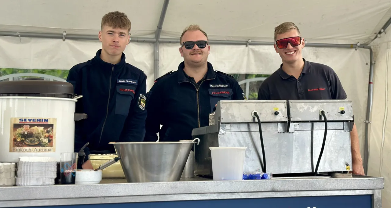Drei Feuerwehrleute in Uniform stehen hinter einem mobilen Getränkeausgabestand mit Kaffeemaschine und Zapfhahn.