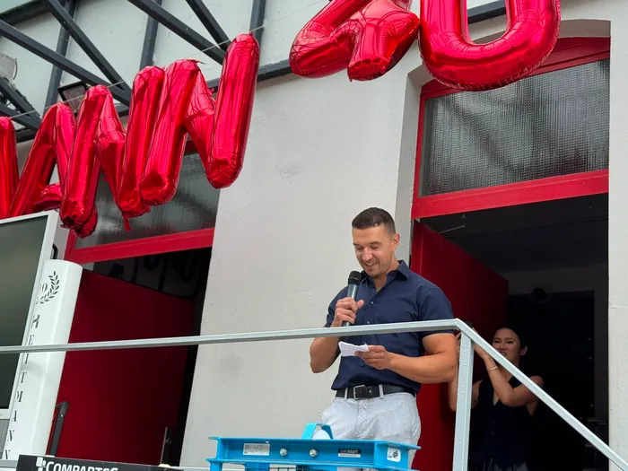 Ein Mann spricht mit einem Mikrofon und einem Zettel in der Hand vor großen roten Luftballons bei einer Jubiläumsfeier.