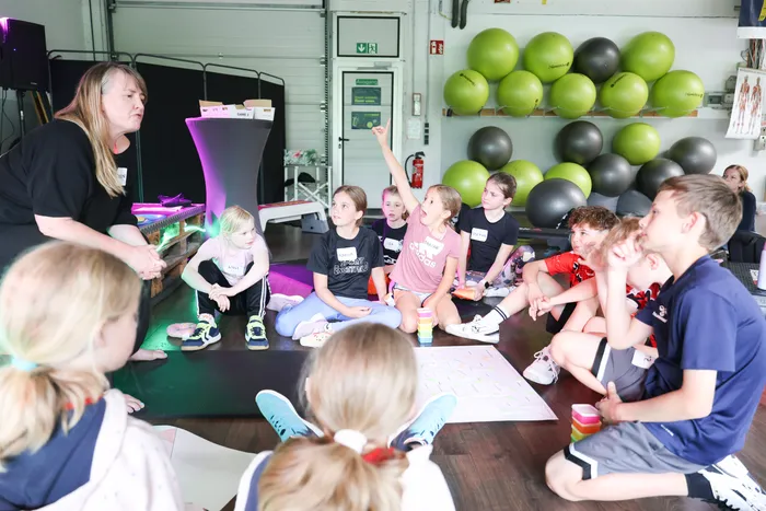 Eine Frau steht vor einer Gruppe von Kindern, die im Kreis sitzen. Einige Kinder heben die Hand, während die Frau spricht. Im Hintergrund sind Gymnastikbälle an der Wand zu sehen.