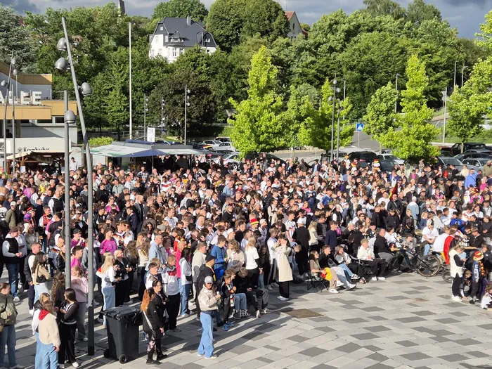 Große Menschenmenge auf dem Otto-Fuchs-Platz jubelt während eines Public Viewings, vermutlich beim Fußballspiel Deutschland gegen Curaçao. Im Hintergrund sind Gebäude und Bäume zu sehen.