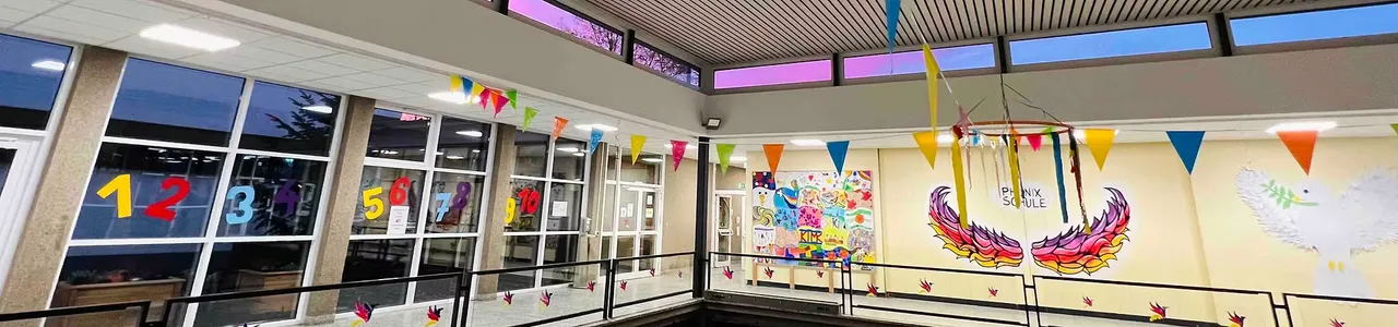 Innenansicht einer hellen Schulaula mit bunten Flaggen und einem Wandbild eines Phönix, der aus den Flammen aufsteigt.