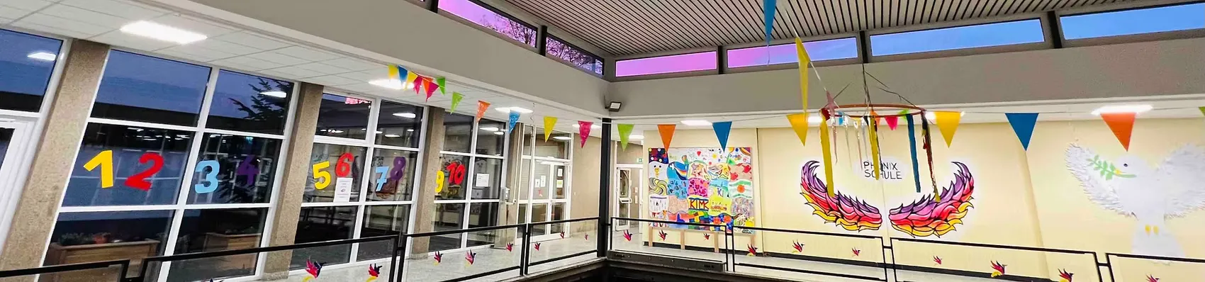 Innenansicht einer hellen Schulaula mit bunten Flaggen und einem Wandbild eines Phönix, der aus den Flammen aufsteigt.