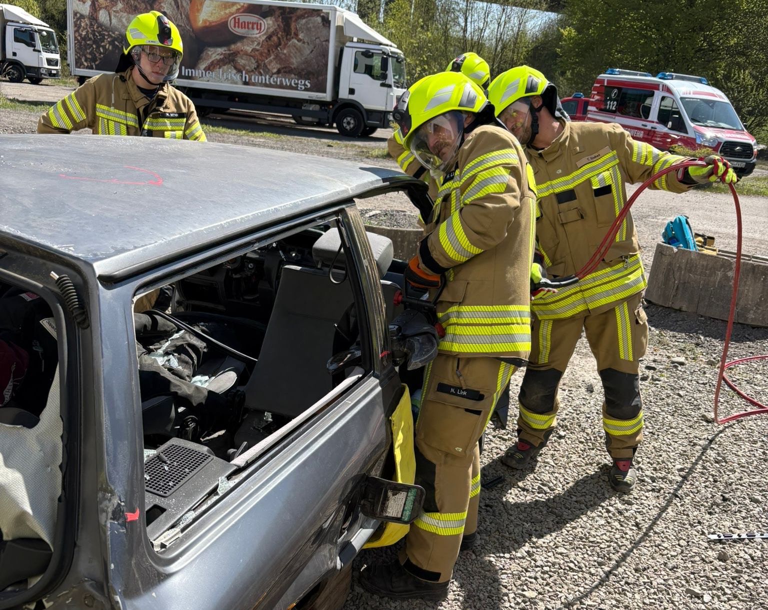 Drei Feuerwehrleute in voller Montur befreien eine Person aus einem stark beschädigten silbernen Auto. Im Hintergrund ein weiterer Einsatzwagen und weitere Einsatzkräfte.