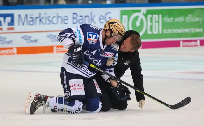 Ein Eishockeyspieler in blau-weißer Uniform kniet auf dem Eis, während ein Mann in schwarzer Kleidung zu ihm hinunterbeugt.