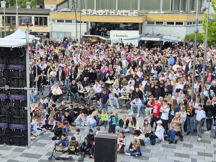 Große Menschenmenge auf dem Otto-Fuchs-Platz jubelt während eines Public Viewings, viele tragen Fußballtrikots. Im Hintergrund ist die Stadthalle zu sehen.