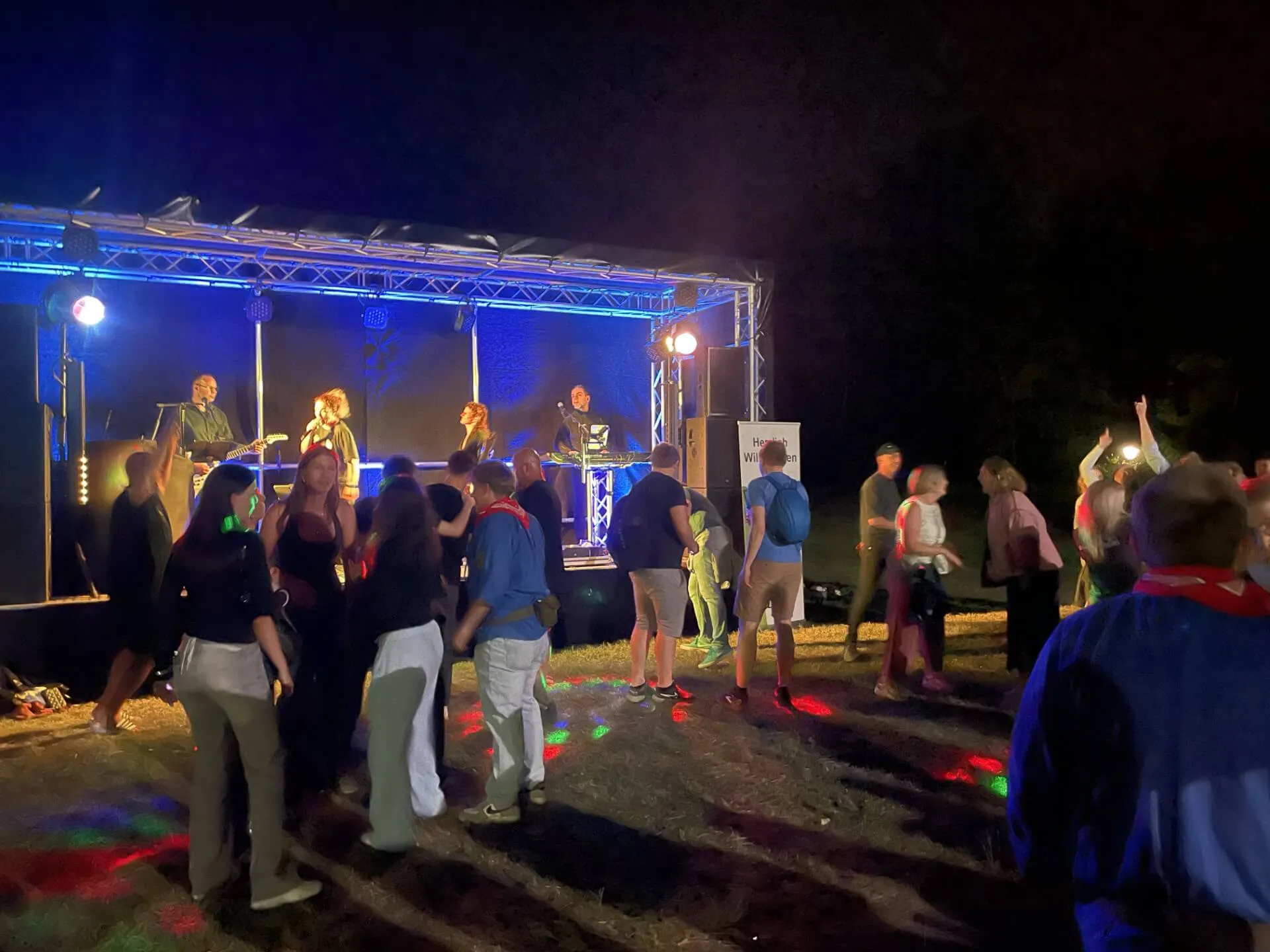 Eine Band spielt auf einer beleuchteten Bühne vor einem Publikum bei einer Feier im Freien. Im Hintergrund sind Bäume und ein Banner mit der Aufschrift „Heinrich Wulffen“ zu sehen.