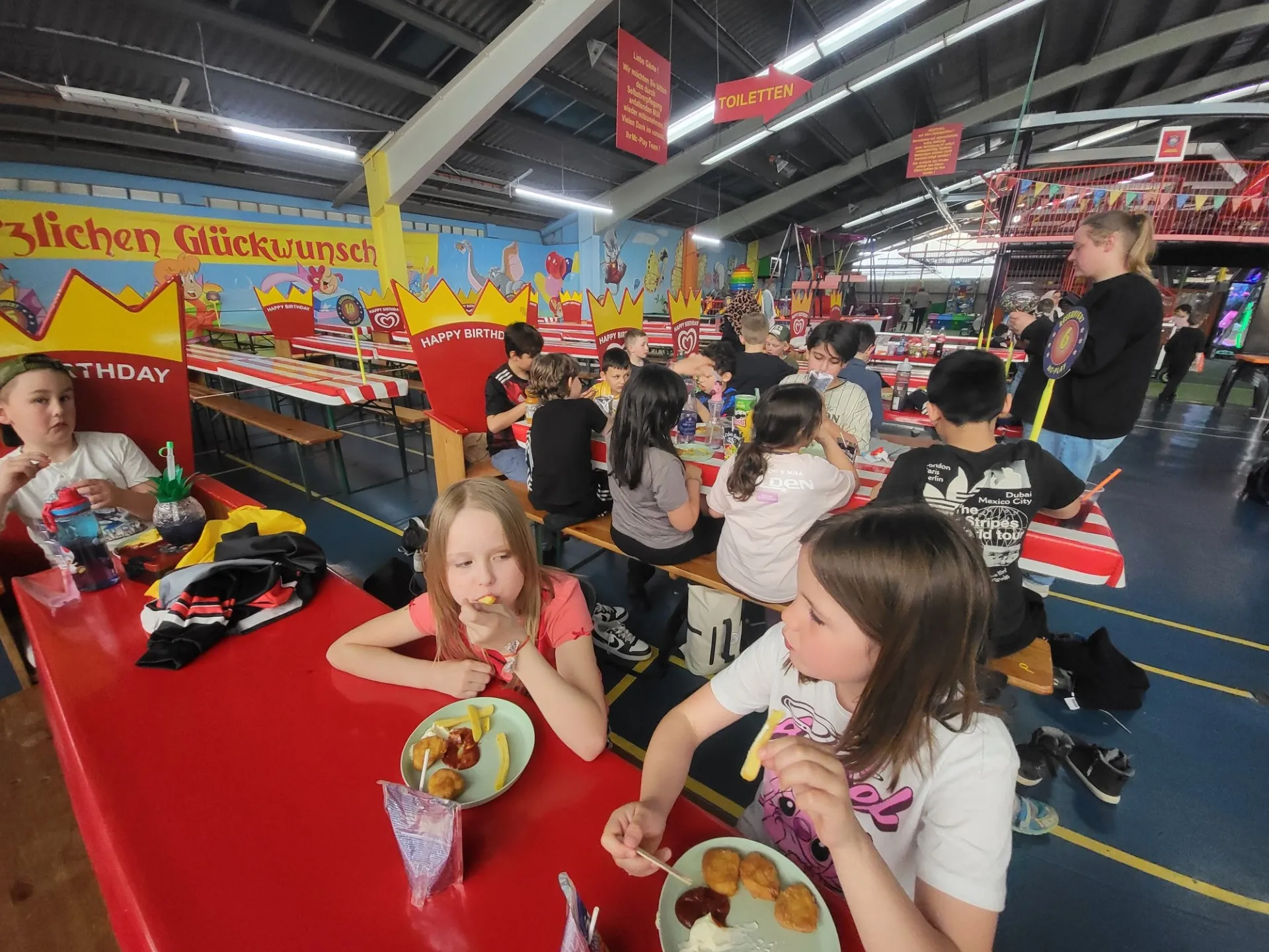 Kinder sitzen an einem roten Tisch und essen Pommes und Nuggets. Im Hintergrund sind weitere Kinder und Jugendliche in einer bunten Halle zu sehen, mit Girlanden und Hinweisschildern.