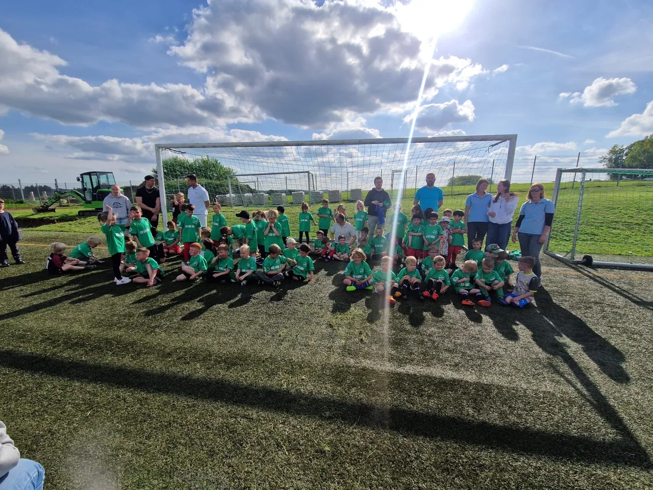 Eine Gruppe von Kindern in grünen Trikots posiert vor einem Fußballtor auf einem Kunstrasenplatz, umgeben von Erwachsenen.