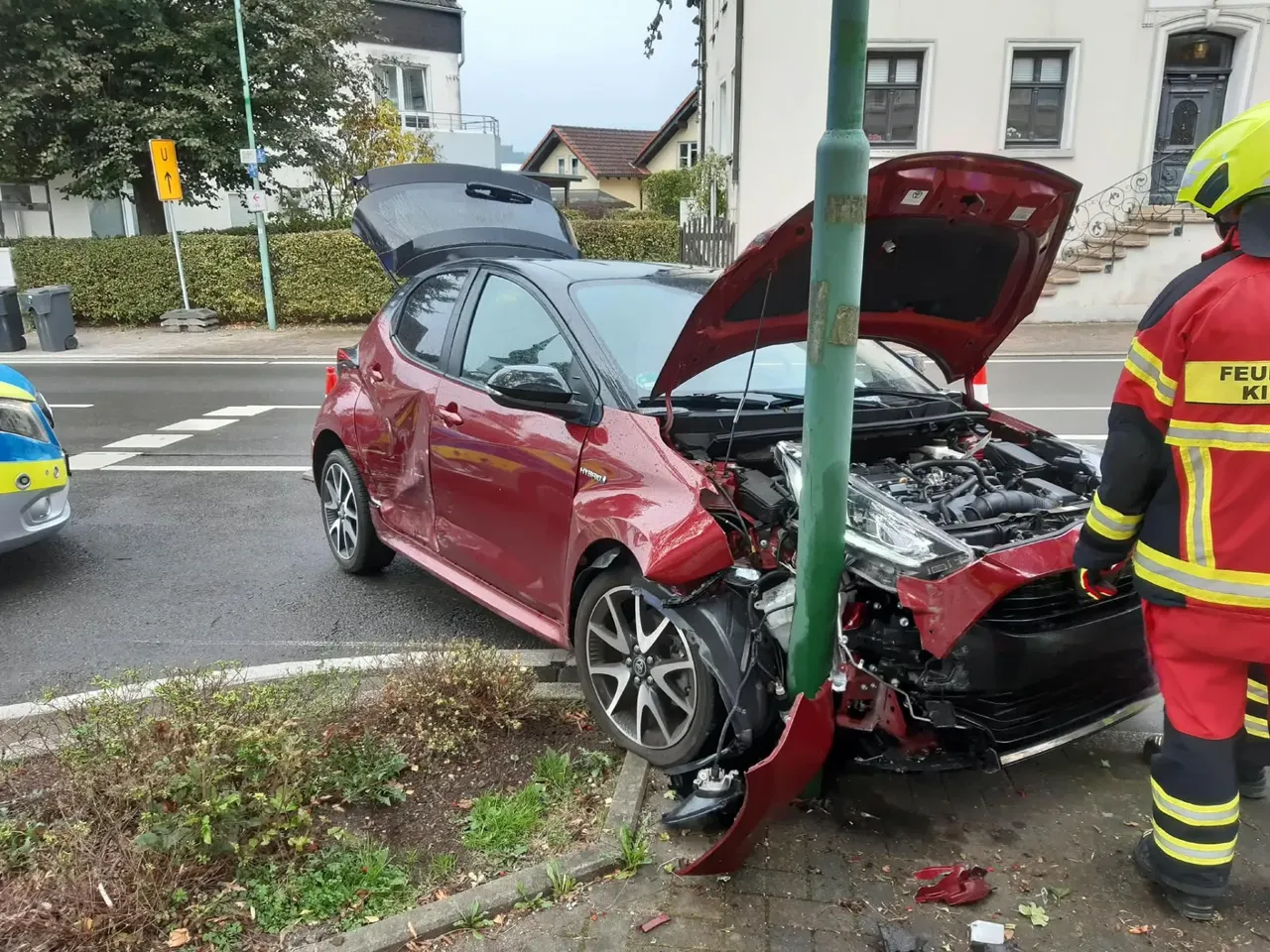 Ein rotes Auto mit schwer beschädigter Front nach einem Zusammenstoß mit einem grünen Pfosten; ein Feuerwehrmann steht daneben.