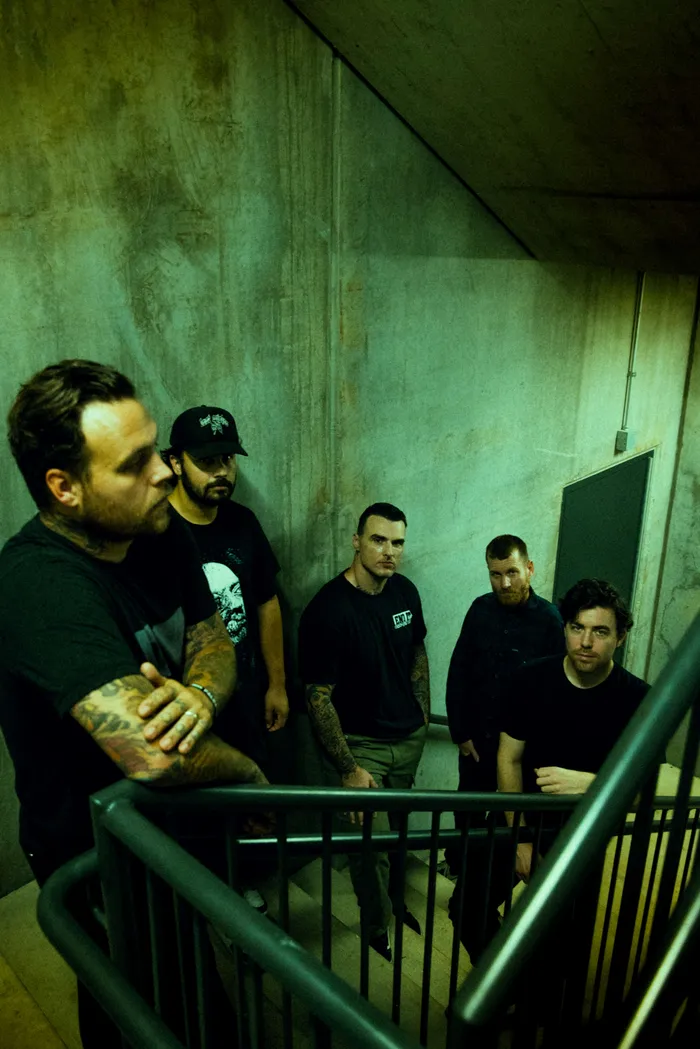 Fünf Mitglieder der Band Stick To Your Guns stehen in einem grünlich beleuchteten Treppenhaus mit Betonwänden.