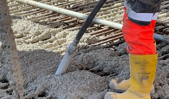 Ein Arbeiter in orangefarbener Hose und gelben Gummistiefeln bearbeitet frischen Beton auf einer Bewehrung aus Stahlstäben.