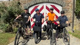 Vier Personen mit Fahrrädern posieren vor einem rot-weiß gestreiften Tor an einem Natursteingebäude.