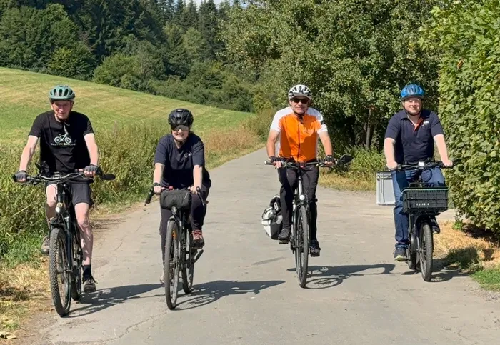 Die Tourguides Bernd Henrichs, Nadine Walter, Ralf Schiwotz und Markus Stumpf fahren mit ihren Fahrrädern auf einem Weg durch eine Landschaft mit Wiesen und Wald.