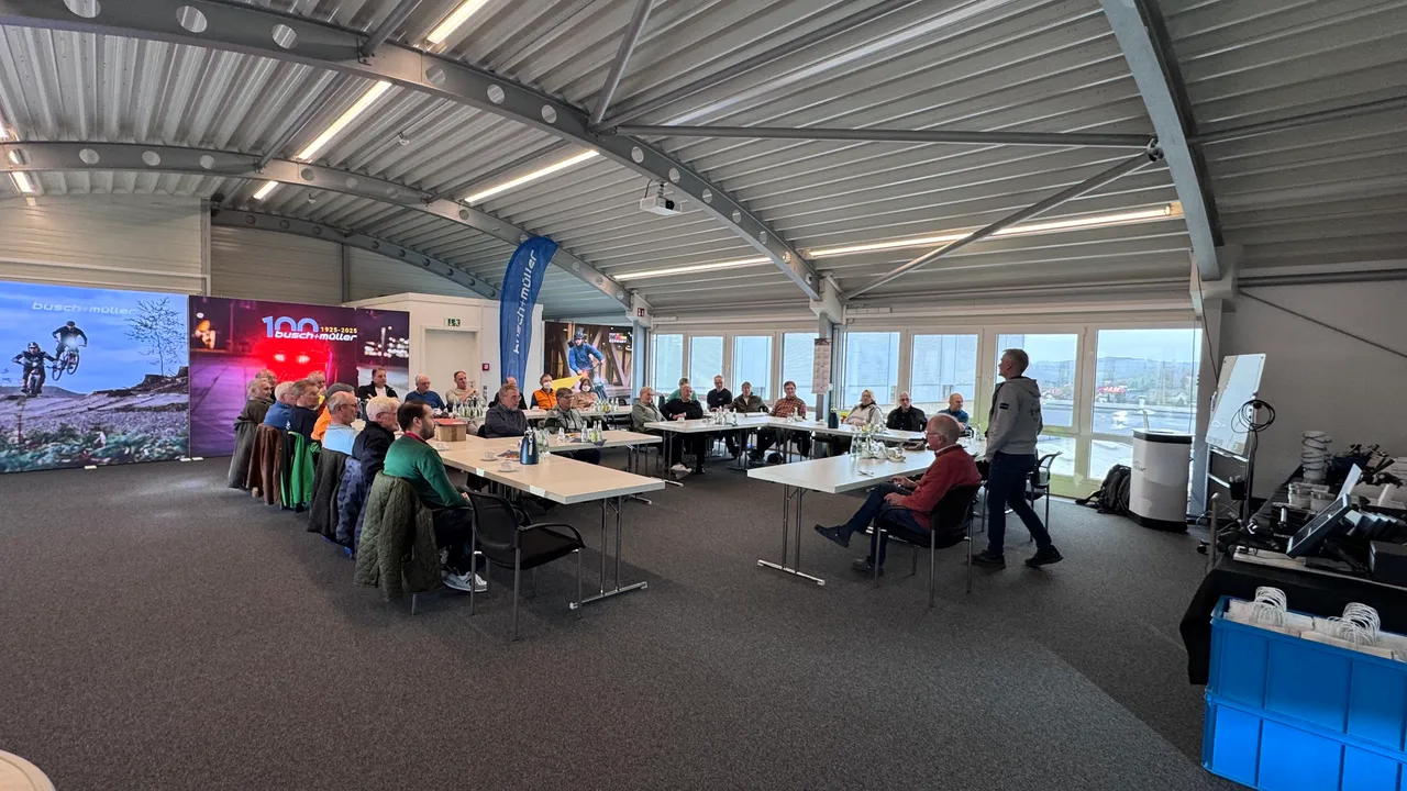 Eine Gruppe von Menschen sitzt an Tischen in einer hellen Halle, während ein Mann im Gespräch steht. Im Hintergrund sind Werbebanner von Busch + Müller zu sehen.