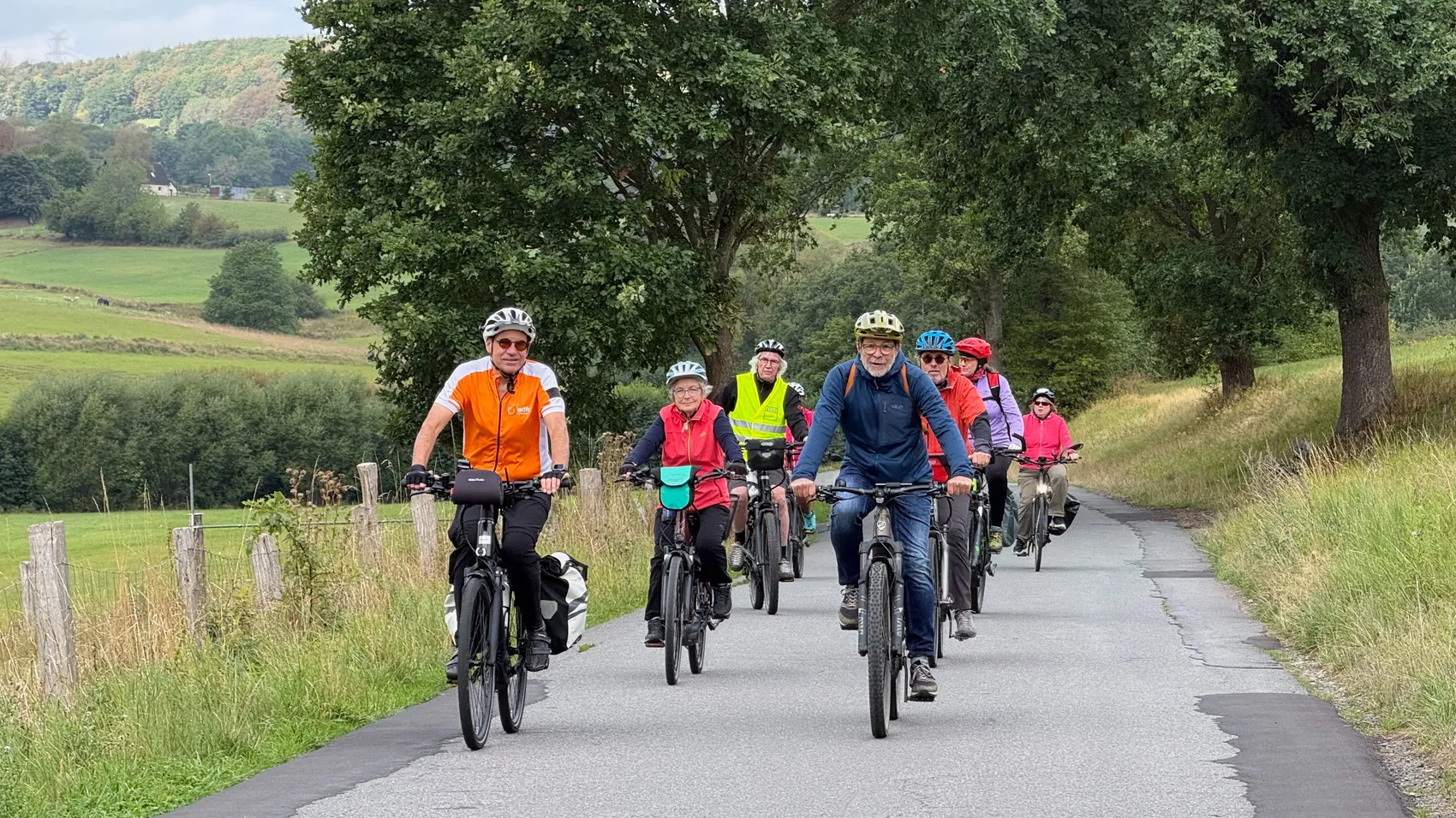 Eine Gruppe von Radfahrern fährt auf einem asphaltierten Weg durch eine grüne Hügellandschaft.