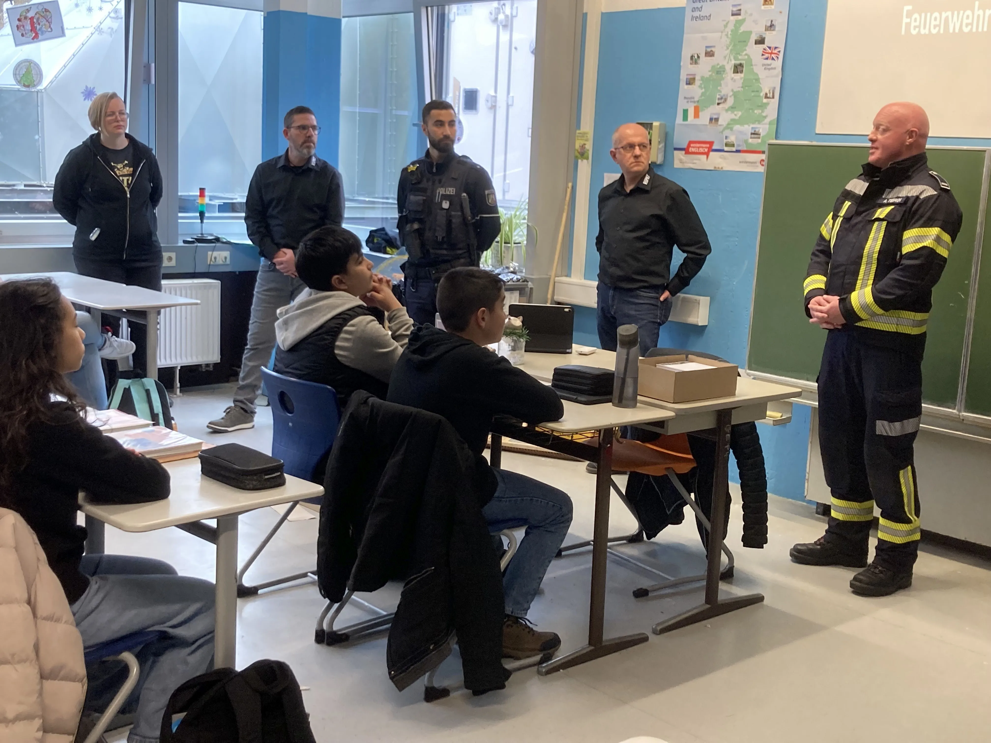 Polizisten und ein Feuerwehrmann sprechen in einem Klassenzimmer mit Schülern über die Arbeit der Soko Respekt. Im Hintergrund eine Karte von Irland und Großbritannien sowie ein Hinweis auf die Feuerwehr.