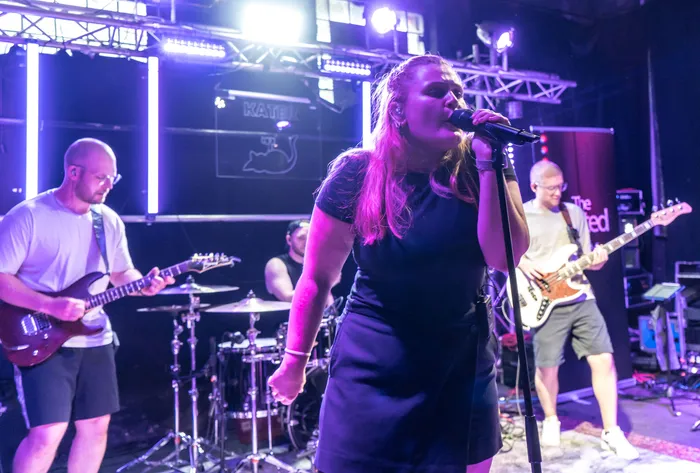 Eine Frau singt in ein Mikrofon, während eine Band im Hintergrund spielt. Das Bühnenbild zeigt den Schriftzug 