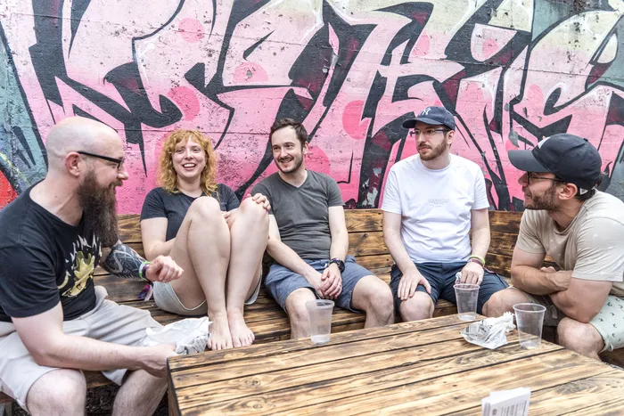 Fünf junge Erwachsene sitzen an einem Holztisch vor einer mit Graffiti besprühten Wand. Sie unterhalten sich und haben Getränke vor sich stehen.