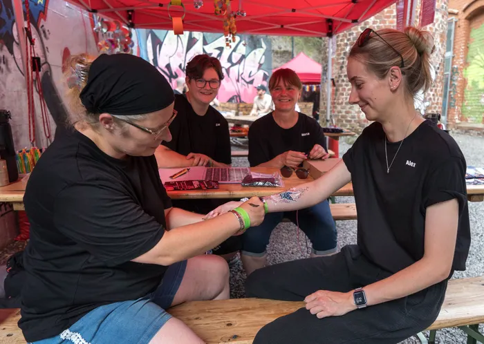 Eine Person trägt ein Tattoo auf den Arm einer anderen Person auf, während zwei weitere Personen im Hintergrund sitzen. Das Festival findet im Freien unter einem roten Zelt statt.