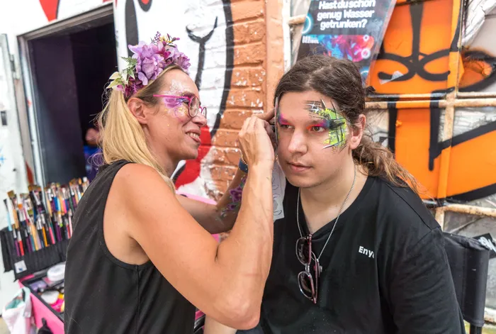 Eine Frau bemalt das Gesicht eines jungen Mannes auf einem Festival. Im Hintergrund ist eine bunte Graffiti-Wand zu sehen.