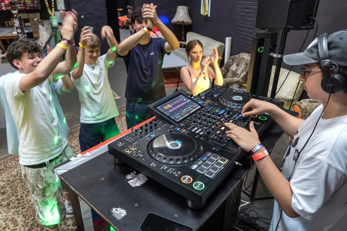 Ein DJ spielt vor einer tanzenden Gruppe junger Leute auf einem Festival im AJZ. Im Hintergrund sitzt eine Person auf einem Sofa.