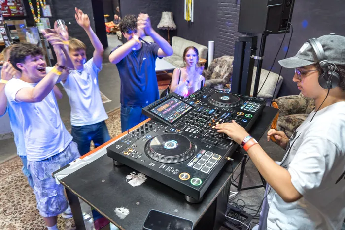 Ein DJ steht hinter einem Mischpult und legt Musik auf, während mehrere Jugendliche im Hintergrund tanzen und die Hände in die Luft heben.
