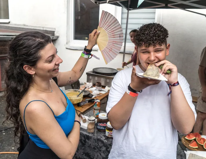 Ein junger Mann mit lockigem Haar beißt in einen Crêpe, während eine junge Frau mit langen, dunklen Haaren ihn mit einem Fächer fächelt. Im Hintergrund steht ein mobiler Crêpes-Stand mit verschiedenen Belägen.