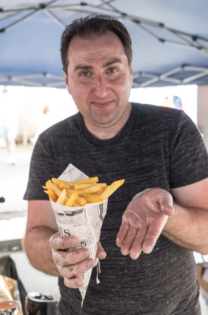 Ein Mann mit dunklem Haar hält eine Papiertüte mit Pommes Frites in der Hand und bietet sie der Kamera an. Im Hintergrund ist ein Zelt und ein Festivalgelände zu sehen.
