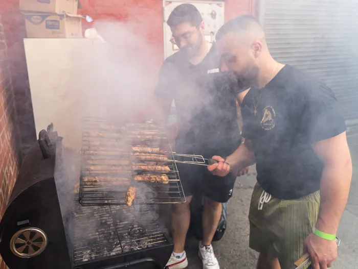Zwei Männer grillen Fleischspieße auf einem Holzkohlegrill im Freien, Rauch steigt auf. Im Hintergrund sind Kartons und weitere Personen zu sehen.