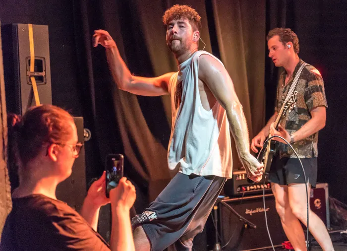 Ein Sänger mit gelocktem Haar und Tanktop performt auf einer Bühne, während ein Gitarrist im Hintergrund spielt. Im Vordergrund filmt eine Person mit einem Smartphone das Konzert.