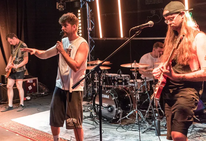 Eine Band spielt auf einer Bühne vor Publikum. Ein Sänger mit lockigem Haar und Shorts steht mit Mikrofon in der Mitte, während ein Gitarrist rechts und ein Keyboarder links von ihm stehen. Im Hintergrund ist ein Schlagzeugset zu sehen.
