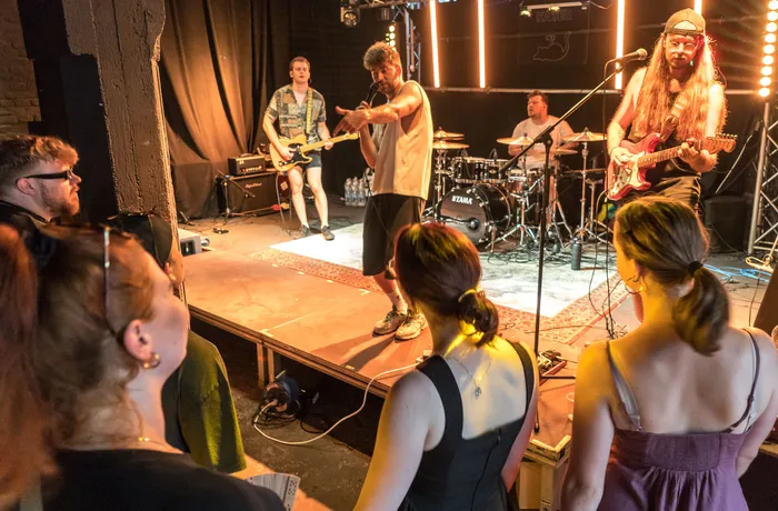 Eine Band spielt auf einer Bühne vor einem Publikum. Ein Gitarrist, ein Schlagzeuger und ein Sänger sind zu sehen, während Menschen im Vordergrund stehen und zusehen.