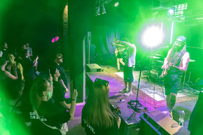 Eine Band spielt vor einem Publikum in einem dunklen Club. Ein Sänger mit Mütze und ein Gitarrist mit langen Haaren stehen auf einer Bühne, während das Publikum mit Smartphones filmt.