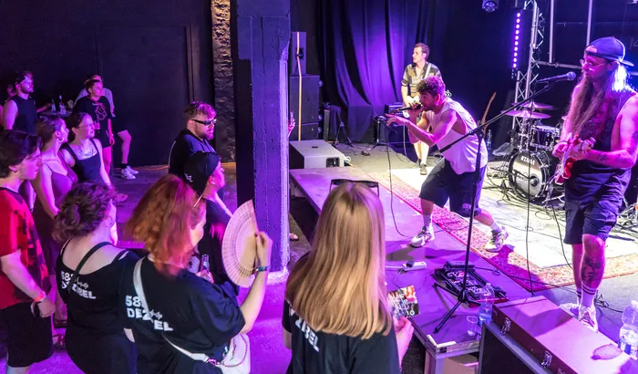 Eine Band spielt vor einem Publikum in einem dunklen Club. Im Vordergrund tanzen und stehen Menschen, einige mit T-Shirts mit Aufschrift. Die Band besteht aus drei Personen, die Instrumente spielen.