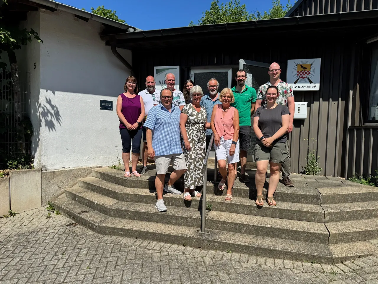 Eine Gruppe von zehn Personen steht vor dem Gebäude des ASV Kierspe, dem Schießsportzentrum am Amselweg. Sie lächeln in die Kamera und bilden eine Gemeinschaft von Vereinsmitgliedern, die an der passiven Schützenmeisterschaft teilgenommen haben.