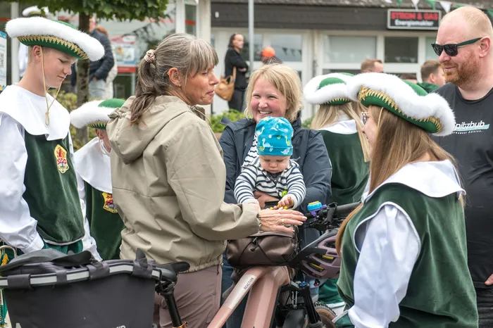 Menschen in grünen und weißen Schützenuniformen mit Federhüten stehen bei einem Festumzug in Kierspe zusammen mit Passanten und einem Baby.