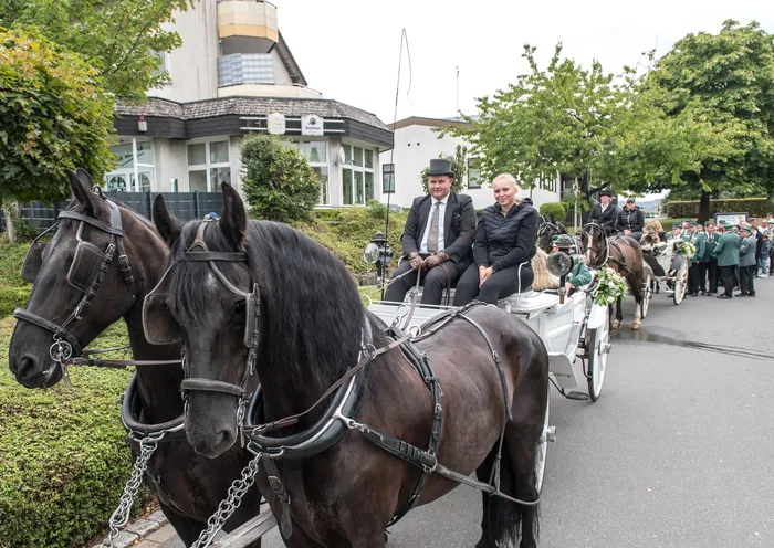 Ein weißer Festwagen, gezogen von zwei schwarzen Pferden, wird von einem Mann mit Zylinder und einer Frau während eines Festumzugs gelenkt.